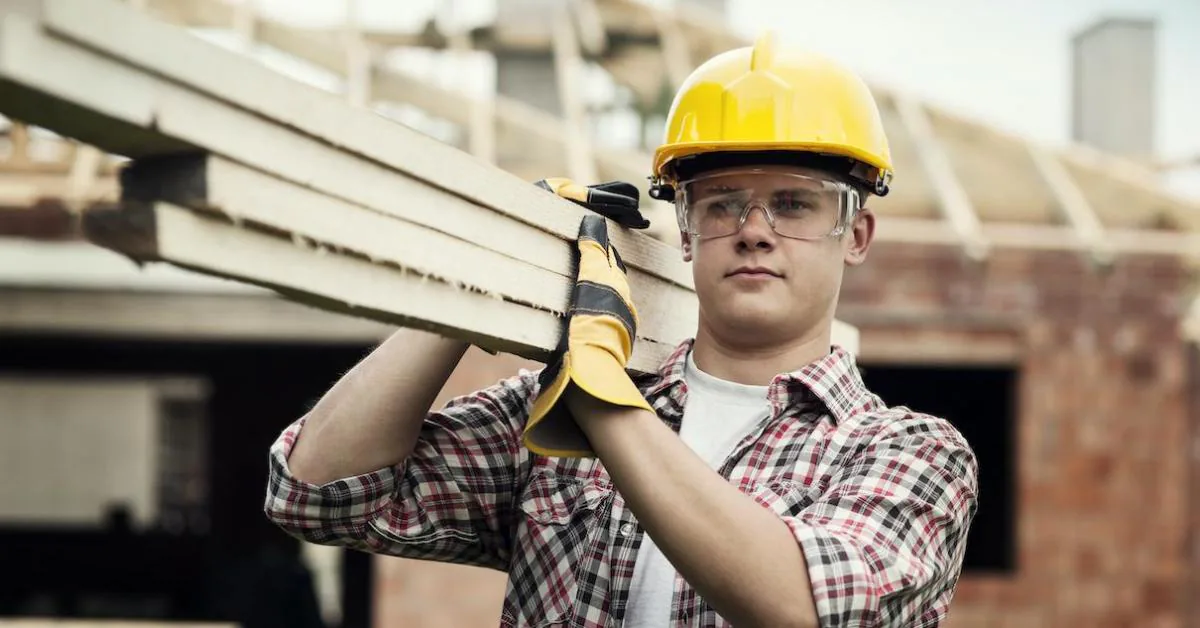 Young labourer in a hard hat carrying timber planks on a UK construction site